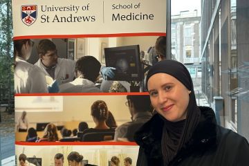 A female undergraduate student wearing a headcovering and dark coat, standing in front of a pop-up banner which shows photographs of medical students and the words School of Medicine